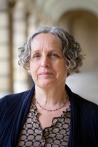 Mary Ann Smart White female with short gray hair and blue eyes wearing a blue blazer and brown blouse standing in a long corridor with arches and pillars.